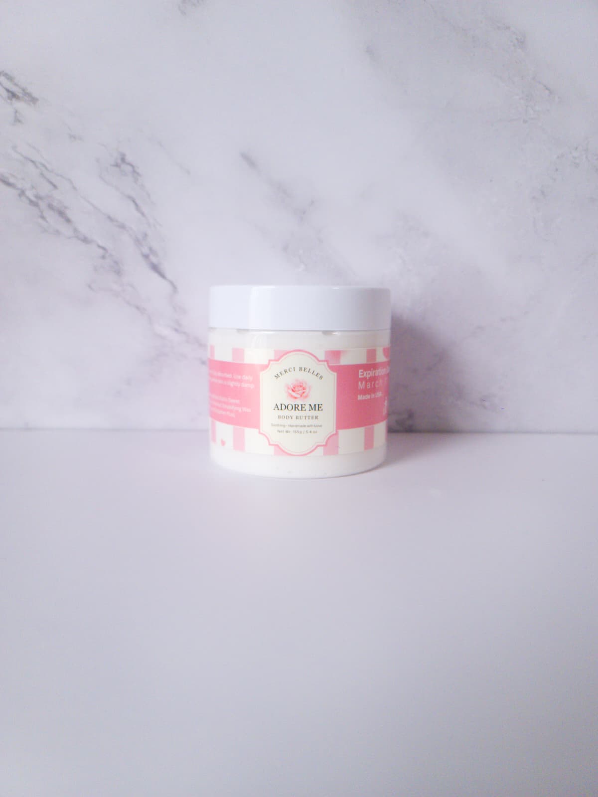 white Body butter in plastic jar with white lid and pink label.
Text: Merci Belles Adore Me Body Butter. Handmade with love. 6oz.